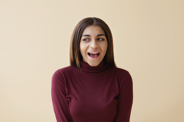 Positive human emotions and feelings. Picture of emotional young mixed race lady looking sideways with excited expression, opening mouth widely, posing against blank copyspace studio wall background