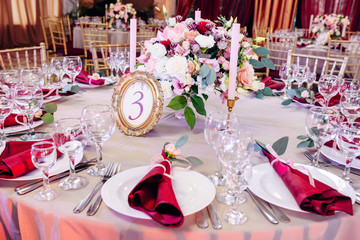 Decorated tables in gold and burgundy colors with plates, knives and forks, candles and glasses, bouquet with flowers and greens on the center