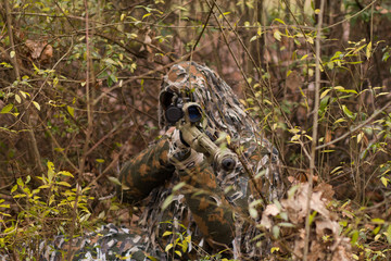 Naklejka na ściany i meble Hunter or soldier hiding in bushes, autumn forest background. Sniper with rifle. 