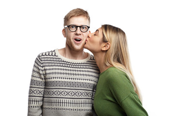Picture of emotional funny young nerdy man wearing rectangular spectacles exclaming excitedly, opening mouth widely as attractive girl kissing him on his cheek. People, love, romance and dating