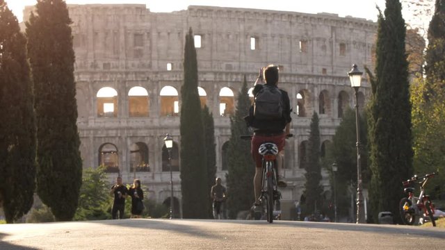 Young man tourist riding bike alone in colle oppio park in front of colosseum on road with trees at sunset in Rome slow motion