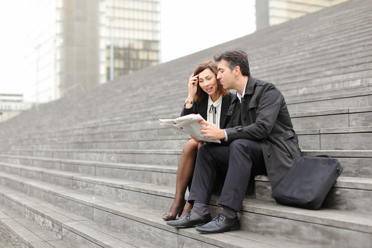 Engineers Male And Female Find Article About Company In Newspaper, Colleagues Sitting On Stairs Reading. Americans Wearing Business Clothes Smiling Communicating. Concept Of Fashionable Cl