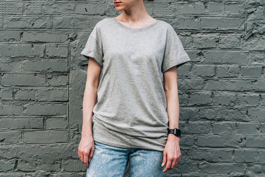 Summer day. Front view. Young millennial woman dressed in gray t-shirt is stands against gray brick wall. Mock up. Space for logo, text, image.