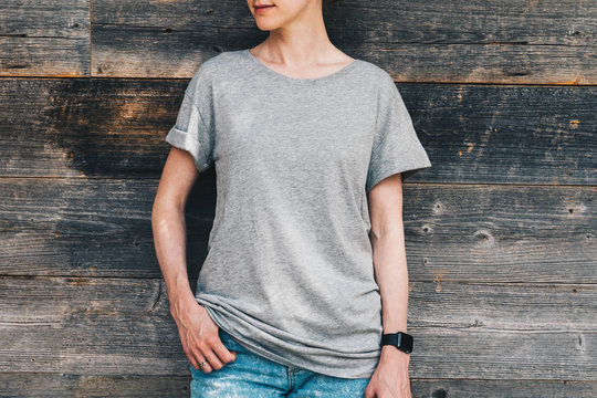 Summer day. Front view. Young millennial woman dressed in gray t-shirt is stands against gray wood wall. Mock up. Space for logo, text, image.