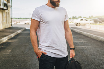 Summer day. Front view. Young bearded millennial man dressed in white t-shirt is stands on city...
