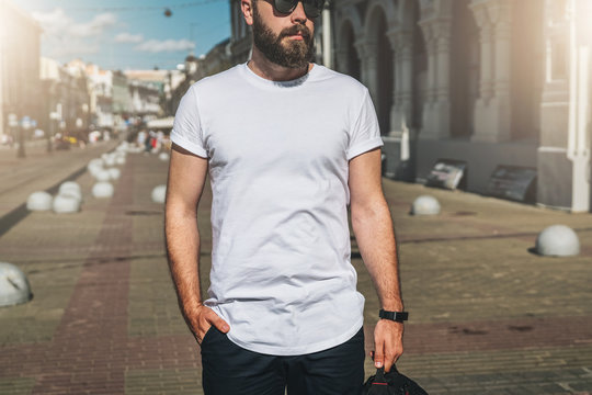 Summer day. Front view. Young bearded millennial man dressed in white t-shirt and sunglasses is stands on city street. Mock up. Space for logo, text, image. Instagram filter, film effect, bokeh effect
