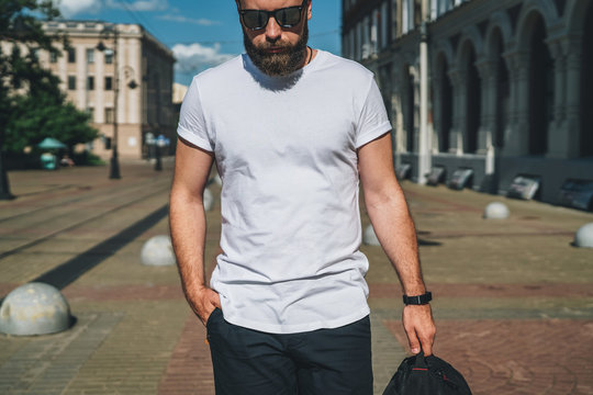 Summer day. Front view. Young bearded millennial man dressed in white t-shirt and sunglasses is stands on city street. Mock up. Space for logo, text, image. Instagram filter, film effect, bokeh effect