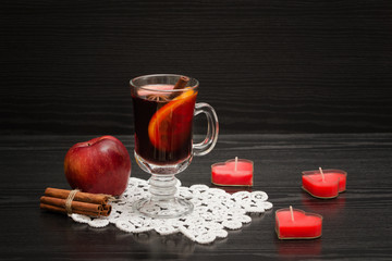 Mulled wine with spices on a lace napkin. Candles in the shape of a heart, cinnamon sticks and apple. Black wood background