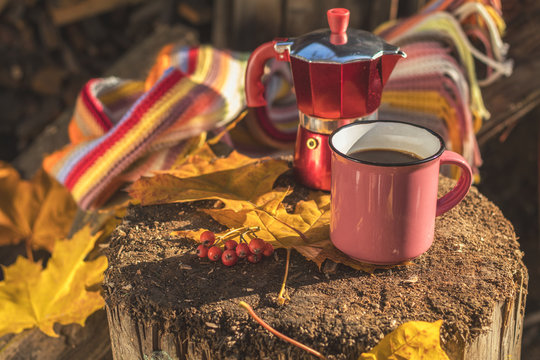 Multicolored scarf, cup of coffee, red coffee maker, yellow maple and oak leaves on the wooden board. Bright autumn background. Sunlight, copy space.