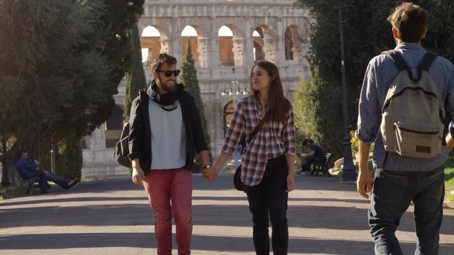 Young couple walks in park road holding hands when attractive man glances at girlfriend. She gets distracted and boyfriend gets jealous. Disoloyal girl and envy. Colosseum in the background in rome at