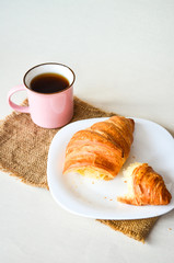 Close up of breakfast, cup of coffee and big tasty croissant on white plate on white table.
