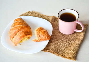 Close up of breakfast, cup of coffee and big tasty croissant on white plate on white table.