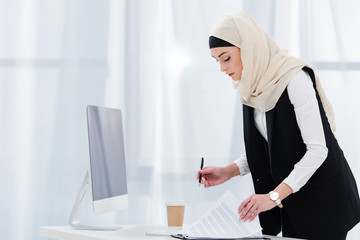 Obraz na płótnie Canvas side view of muslim businesswoman doing paperwork at workplace in office