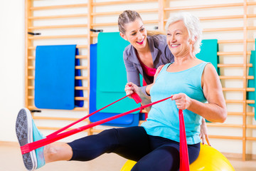 Senior woman with stretch band in fitness gym being coached by personal trainer
