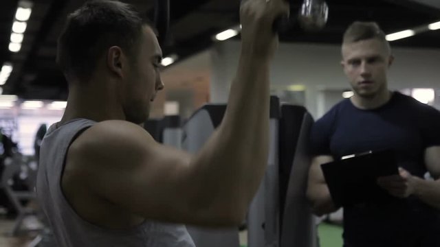 Young bodybuilder is doing wide grip pulldowns exercise with coach in gym. Handsome man trains intensively in modern sports club with instructor. Hardener person in sportswear makes effective practice