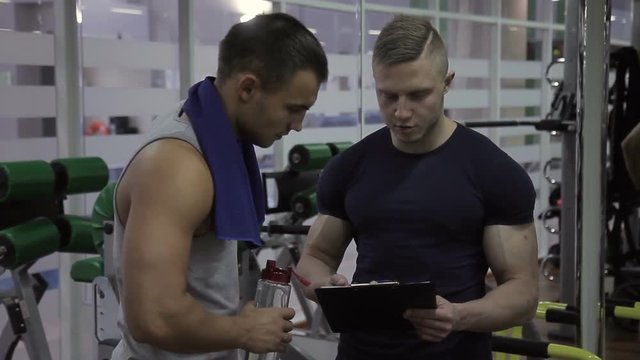 In the gym athlete and coach look at results of training on the sheet. Two men discuss progress achieved during exercise near iron Can athlete with a towel around his neck drinking water.