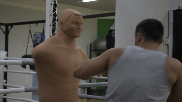 Young man is beating boxing mannequin in modern sports club. Athlete makes strikes with fists on professional equipment. Handsome guy is trained intensively, using rubber punching bag in form of human