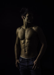 Fototapeta premium Handsome, fit muscular young man shirtless, wearing only jeans standing on black background, looking away to a side