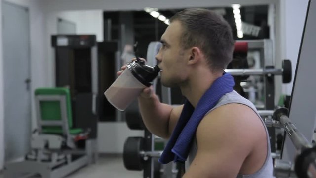 Young man drinks protein cocktail in gym indoors. Promising athlete takes sip of chocolate whey beverage from shaker in break between exercises. Person with towel around his neck spends good time in