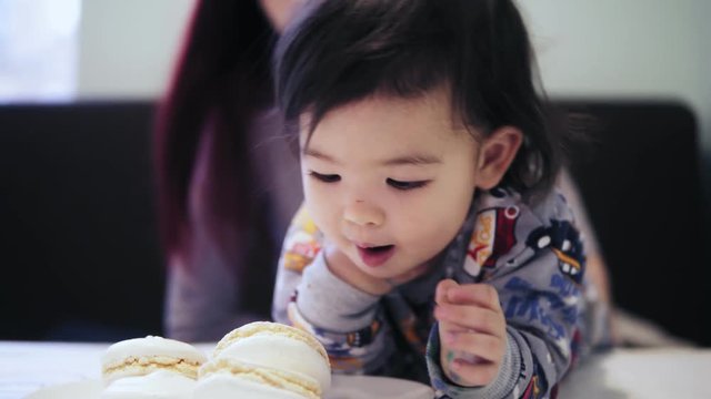 An adorable cute asian toddler boy enjoys eating macaroons with his mother sitting beside him.