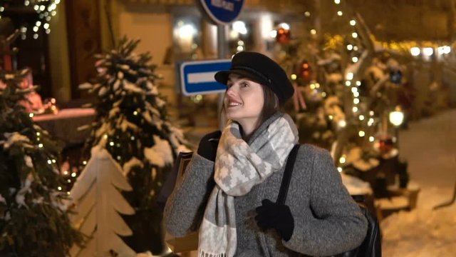 Relaxed girl in gray coat and black cap walking through the night city with shopping bags, slowmotion of snowtime enjoyment