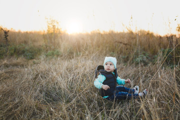 Naklejka na ściany i meble Little cute child baby boy in turquoise hat, vest, jeans sitting on dry high grass background on spring or fall nature. Little kid son. Parenthood family day 15 of may love parents children concept.