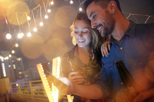 Couple having a drink at rooftop party and using smartphone