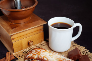 A cup of coffee with a cake, with chocolate sweets and an old hand grinder on a black textured table.