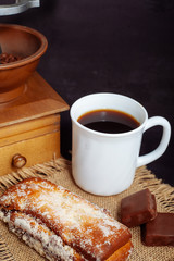 A cup of coffee with a cake, with chocolate sweets and an old hand grinder on a black textured table.