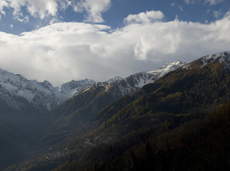 Obraz premium landscape: silhouette of mountains, Antrona Valley, near Swiss, snow on peaks, colored larch forest, sunset, ray of light, Alps, blue sky, clouds, village, late autumn, winter, Piedmont, Italy
