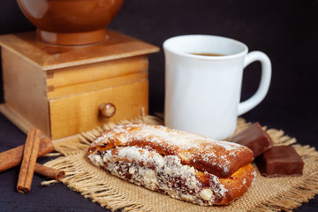 A cup of coffee with a cake, with chocolate sweets and an old hand grinder on a black textured table.