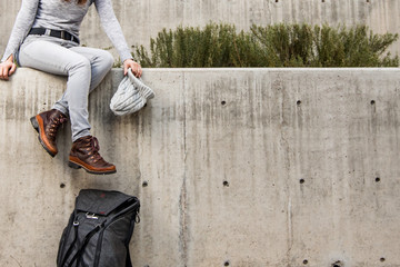 Woman dangling legs off the edge of a concrete ledge with grey beanie hat in hand and  backpack leaning against wall. Lots of negative space to the right. - obrazy, fototapety, plakaty