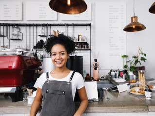Young smiling barista wearing apron, standing at cafe counter - obrazy, fototapety, plakaty