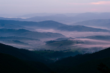 Early morning nature landscape with a view at foggy village in mountains. Scenic panorama with purple toning. Mist falling over conutry houses lost in green hills with forest. Contryside valley.