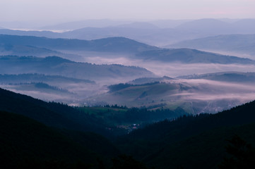 Early morning nature landscape with a view at foggy village in mountains. Scenic panorama with purple toning. Mist falling over conutry houses lost in green hills with forest. Contryside valley.