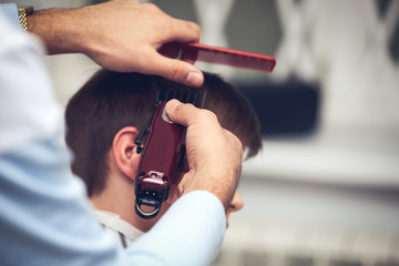 Obraz na płótnie Canvas Barber is doing haircut to Caucasian boy using hair clipper in barbershop.