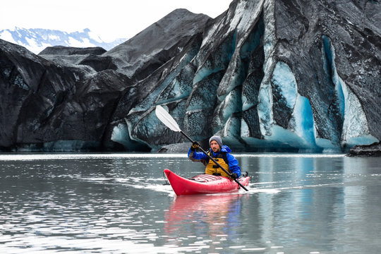 Kayaker Paddling Away From Dark, Moraine Covered Glacier In Alaska