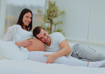 the man leaned against to the baby bump of his pregnant wife, who is lying on the bed.