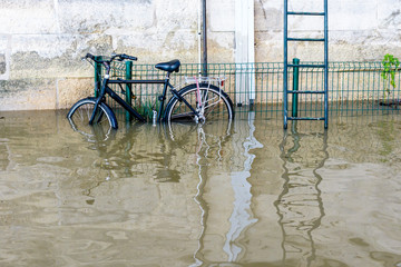 A bicycle attached with a U-lock to a grid, next to a metallic escape ladder, on the riverbanks of the Seine with water at mid-height after the swollen river bursts its banks during a winter flood.