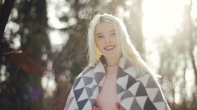 Beautiful young blonde girl with a warm blanket on her shoulders in the winter forest, looking into the camera and smiling