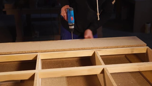 close up shot of the men's hands, who holds the electric drill in order to make holes in the particle board, the carpenter pierces the long board in the workshop