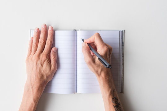 High angle view of woman's hands holding black pen above blank journal on white table