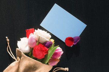 Flower bouquet and message card with petals - Colorful bouquet of diverse flowers wrapped in a jute sack and a blank blue paper note with flower petals on it, on a black background.