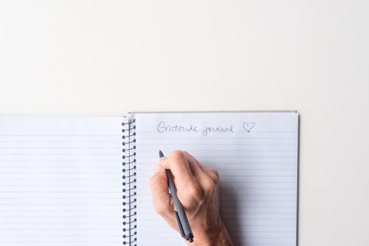 Directly above view of woman's hands holding black pen above spiral notebook gratitude journal on white table.