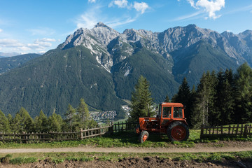 An orange tractor sits on a hill overlooking a town in the Alps. There is an old path and a fence beside the tractor. There are trees behind the tractor and fence line.