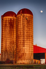 Vertial photo of two silos colored by the sunset with an old vehicle and a red roof with a bright...