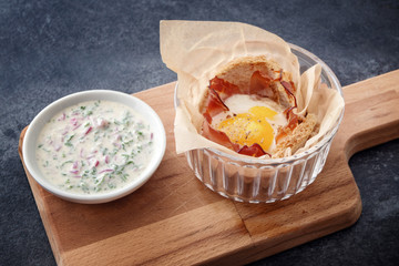 egg muffin with bacon and toasted bread and a fresh cheese dip with herbs on a wooden cutting board, dark slate background
