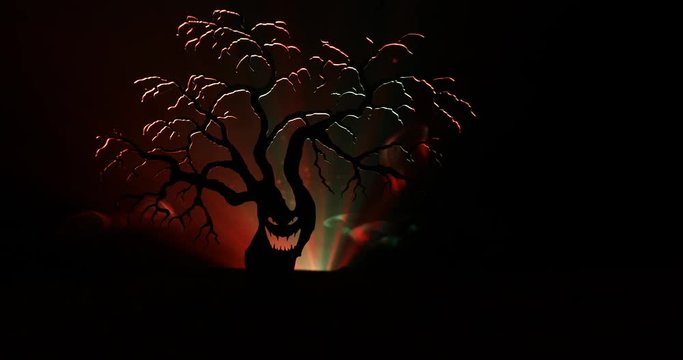Silhouette of scary Halloween tree with horror face on dark foggy toned background with moon on back side. Scary horror tree with zombie and monster demon faces.