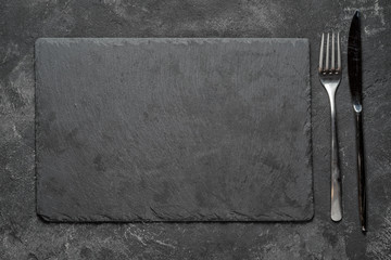 empty black granite stone rectangle board with fork and knife on black textured cement background,...