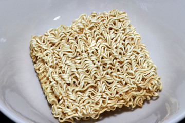 Raw instant noodles squares shape closeup in the white bowl. it is in a precooked and dried noodle block, with flavoring powder and/or seasoning oil.